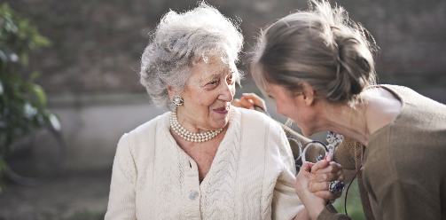 Two women, one elderly and one middle-aged, share a tender moment outdoors with smiles and gentle hand-holding.