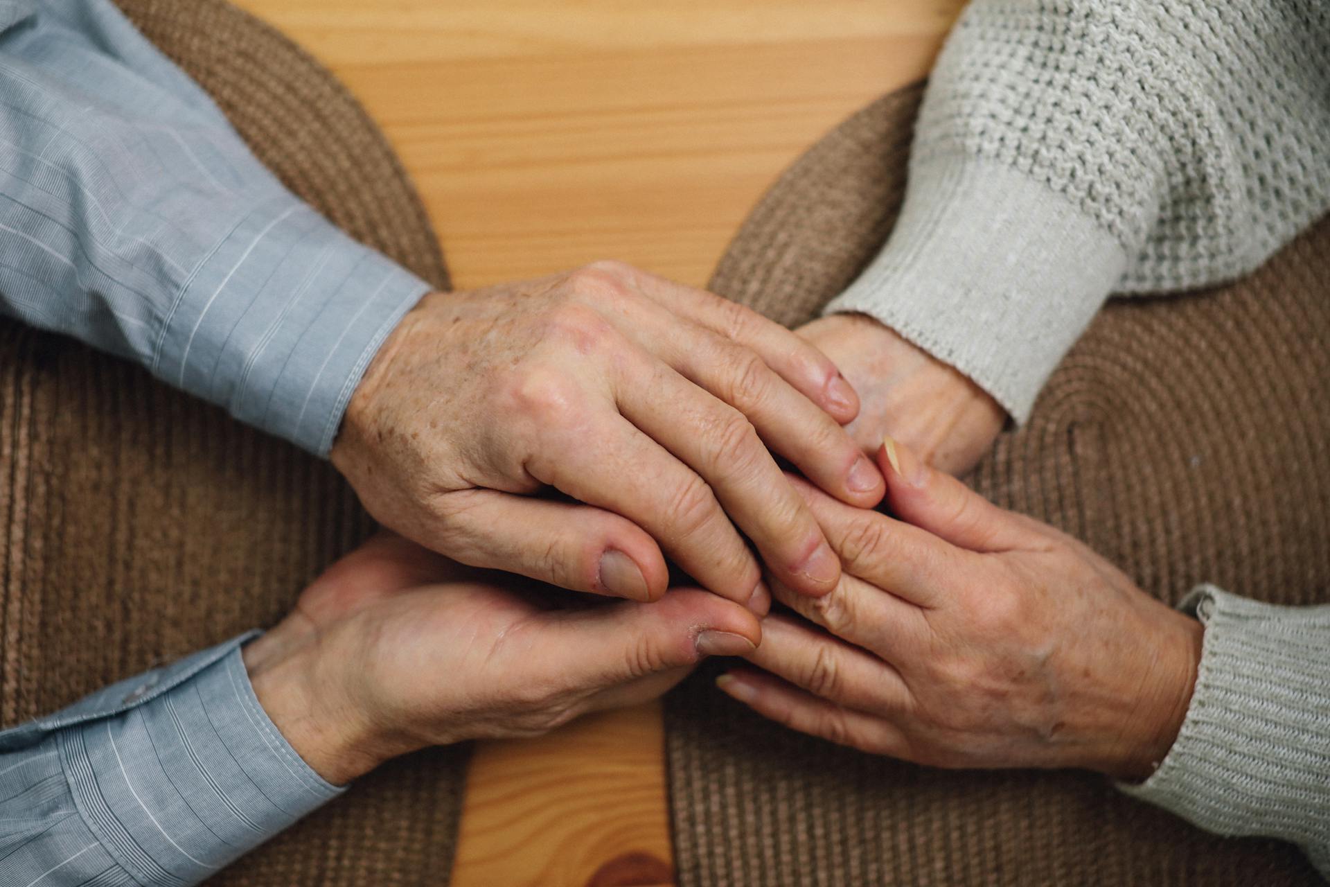 Two pairs of elderly hands gently holding each other on a wooden table with a brown woven placemat.